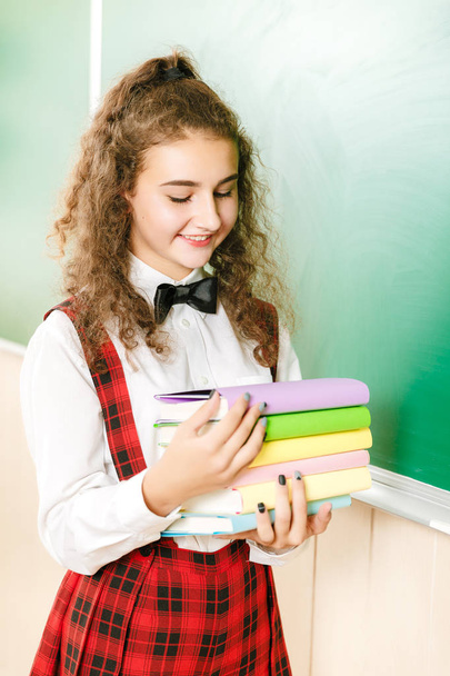 joyeuse écolière en uniforme scolaire debout dans la salle de classe près du tableau noir avec des livres
. - Photo, image