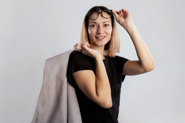 Портрет симпатичной кавказской модели в черной рубашке и больших очках, швыряющей пальто через плечо улыбающейся и одной рукой удерживающей или выпрямляющей очки, стоящие на сером фоне
 - Фото, изображение