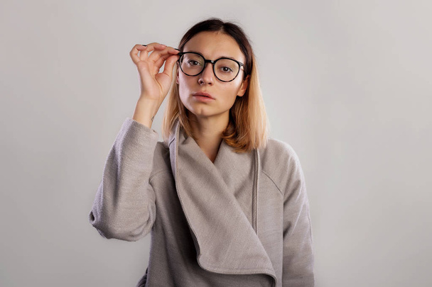 Studio portrait female in big glasses, standing in grey coat and straightens glasses over gray background - Фото, изображение