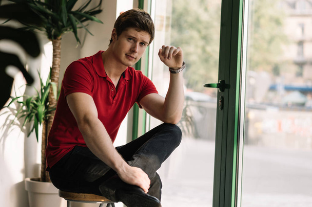 Portrait of young handsome guy wearing red trendy t shirt looking at camera sitting in modern coffee shop. Man fashion model with stylish hairstyle posing for picture indoors.   - Valokuva, kuva