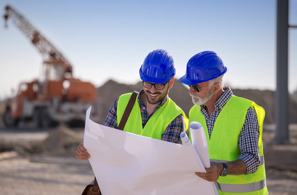 Deux ingénieurs satisfaits regardant les plans sur le chantier avec des machines de construction en arrière-plan
  - Photo, image