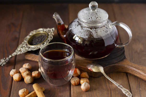 Tea in glass cup with teapot near, vanilla pod and hazelnut at wood background, with spoon and strainer near. - Photo, Image