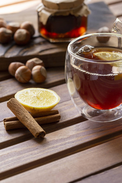 Tea in glass cup with lemon and vanilla pod at wood background. Hazelnut and jam in jar at wood board behind. - Fotó, kép