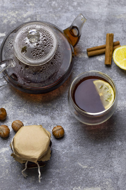 Tea in glass teacup with lemon. Near glass teapot, cinnamon sticks, jam in jar, hazelnuts and lemon at gray background - Photo, Image