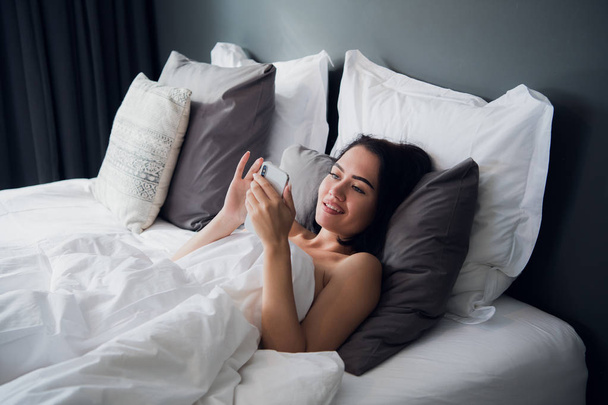 Brunette girl is reading text message from her boyfriend on mobile, while is lying in the bed. Cheerful young woman covered by blanket is watching video message on cell phone before of after sleeping. - Φωτογραφία, εικόνα