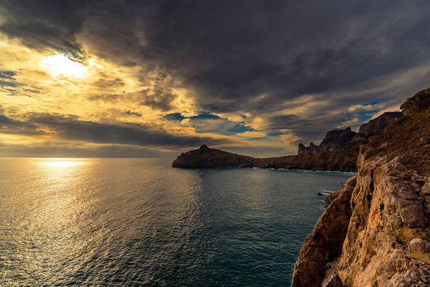 Denizdeki günbatımının panoramik görüntüsü, Cape Kapchik, Golitsyna Patikası, Novy Svet, Sudak, Kırım, Ukrayna. Sağda, güneşte bir kaya parçası, bir deniz, uzakta uzun bir pelerin, parlak renkli bulutlar. - Fotoğraf, Görsel