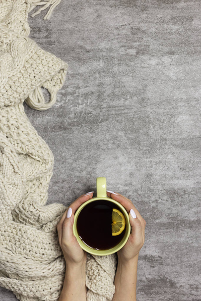 Cup of tea with lemon in hands, knitted scarf near, at gray background - 写真・画像