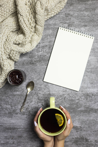 Cup of tea with lemon in hands, knitted scarf, raspberry jam, spoon and notepad near, at gray background - Photo, image