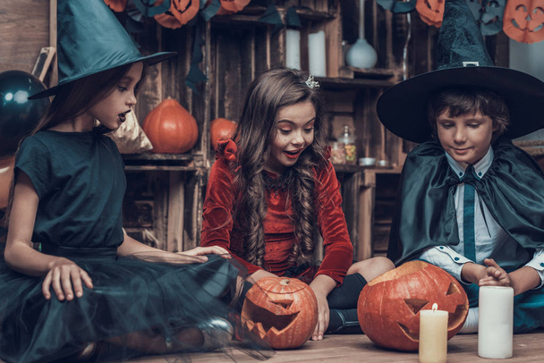 Adorable Little Children in Halloween Costumes. Cute Smiling Kids wearing Scary Halloween Costumes Sitting on Floor next to Carved Pumpkins and Candles. Cute Children Celebrating Halloween - Photo, image
