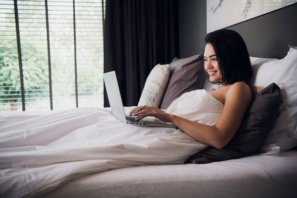 Mujer joven en casa sentada en la cama despertó navegando portátil
 - Foto, Imagen
