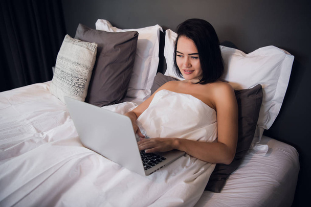Young woman at home sitting on bed woke up browsing laptop - Фото, изображение
