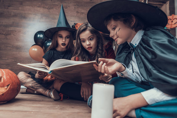 Little Children in Halloween Costumes Reading Book. Cute Smiling Kids wearing Scary Halloween Costumes Sitting on Floor next to Jars full of Candys and Candles. Celebration of Halloween - Photo, image