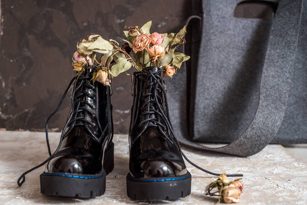 Yellow and pink flowers grow in the old black boots for original decorate. Lacquer black shoes with high soles. Gray felt bag. Concept of warm autumn or winter - Foto, Bild