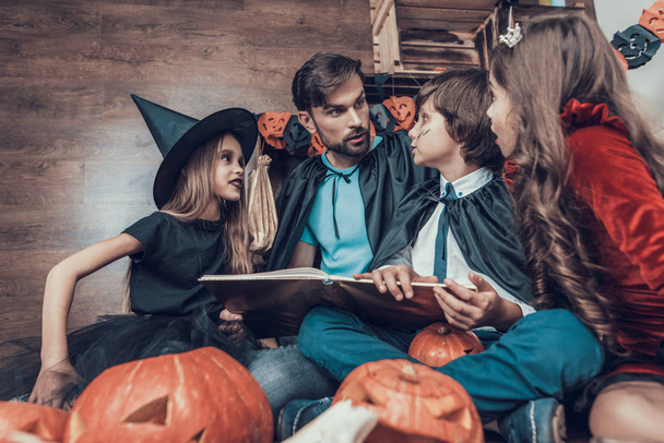 Man and Children in Costumes Reading Book Together. Mignon sourire enfants et homme adulte portant effrayant costumes d'Halloween assis sur le sol à côté de citrouilles sculptées. Célébration d'Halloween
 - Photo, image