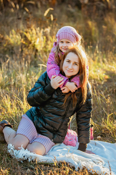 Young beautiful mother with her daughter on a walk on a sunny autumn day. They sit on a plaid on the grass close to each other, the daughter hugs mom from the back and they smile. portrait - Foto, Imagem