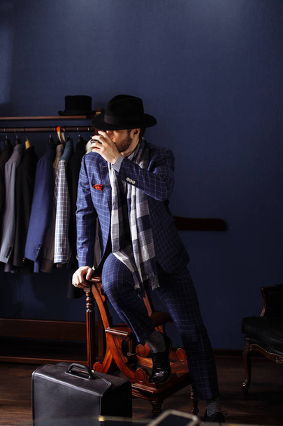Elegant man in fashion suit and hat posing at atelier with beverage - Фото, изображение