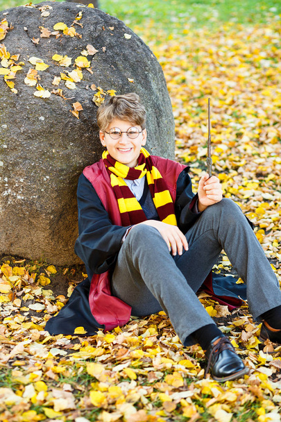 boy in glasses stands in autumn park, holds wand  in his hands, book, wears in black suit and striped scarf.  Halloween costume, cosplay - Photo, Image