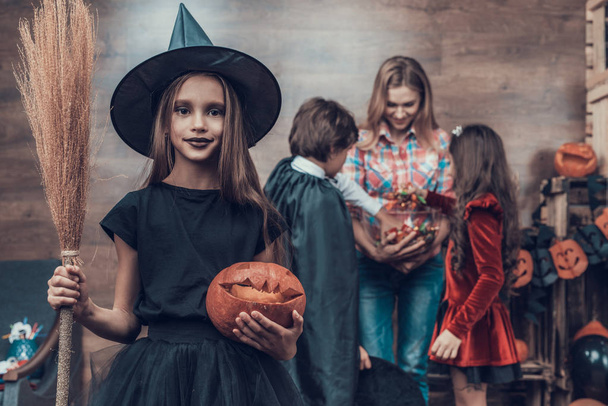 Young Girl wearing Witch Costume Holding Broom. Woman gives Candys to Kids wearing Halloween Costumes. Happy Halloween Party with Children Trick or Treating. Celebration of Halloween - Photo, image