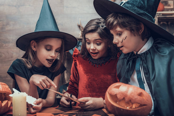 Kids in Costumes Making Halloween Decorations. Adorable Little Children in Costumes Sitting at Table and Cutting Colorful Pieces of Paper with Scissors. Young Kids having Fun Celebrating Halloween - Fotografie, Obrázek