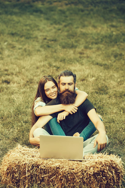 Couple in love, Smiling, adorable girl or cute woman and bearded man, hipster, with beard with long hair using laptop on hay bale on grass on summer day on natural background. Technology, nature - Foto, immagini