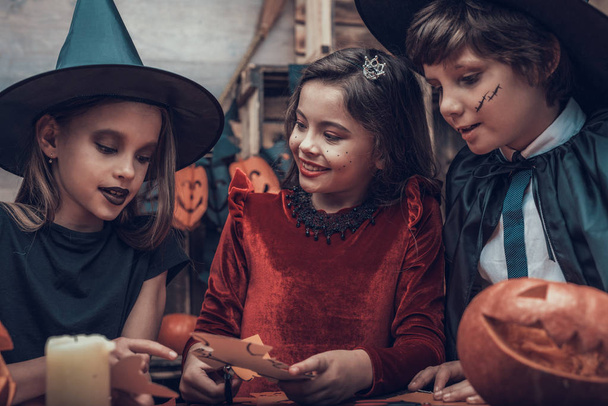 Kids in Costumes Making Halloween Decorations. Adorable Little Children in Costumes Sitting at Table and Cutting Colorful Pieces of Paper with Scissors. Young Kids having Fun Celebrating Halloween - Photo, image