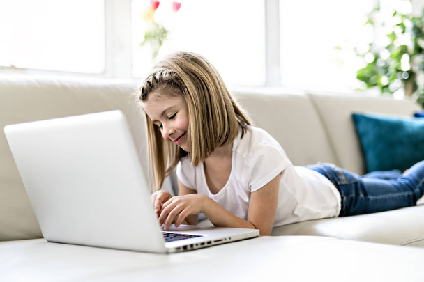 Happy little girl at home working with a laptop on sofa - Photo, image