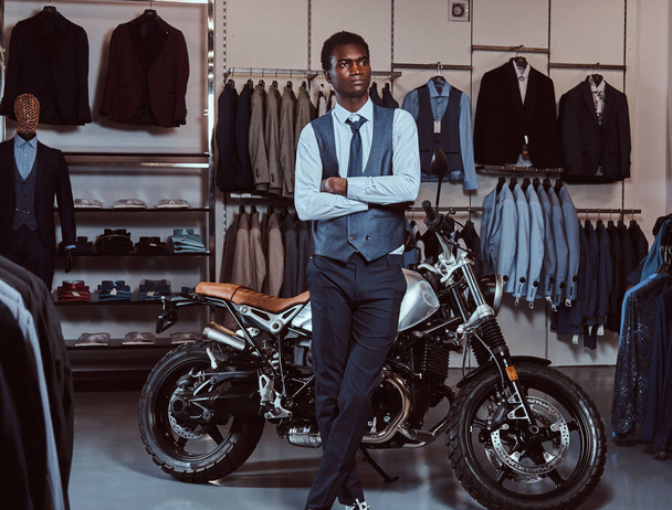 Elegantemente vestido joven afroamericano posando con brazos cruzados cerca de moto deportiva retro en la tienda de ropa para hombres
. - Foto, Imagen