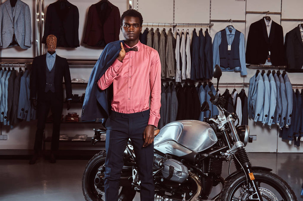 Joven afroamericano vestido con ropa formal elegante posando cerca de la moto deportiva retro en la tienda de ropa para hombre
. - Foto, imagen