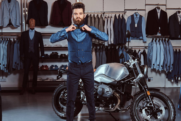 Homme élégant vêtu d'une tenue élégante posant avec les mains sur noeud papillon près de moto de sport rétro au magasin de vêtements pour hommes
. - Photo, image