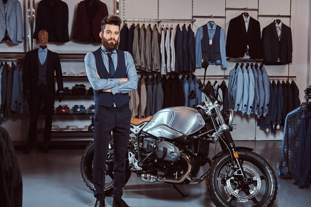 Homme élégamment habillé posant avec les bras croisés près de moto de sport rétro au magasin de vêtements pour hommes
. - Photo, image