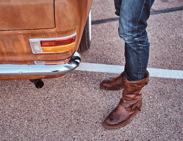 Cropped photo of a male in jeans and boots standing near retro car in the city parking. - Photo, Image