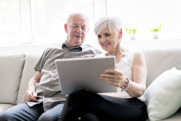 Aanhankelijk aantrekkelijke oudere echtpaar samen zittend op een bank met tablet - Foto, afbeelding