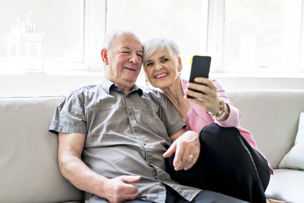 Affectionate attractive elderly couple sitting together on a couch taking selfie - Foto, Imagen