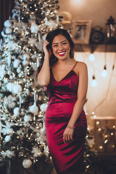 Joven hermosa chica asiática es vestido rojo de moda sentado en casa cerca del árbol de Navidad en el interior acogedor. Interior con decoraciones navideñas
. - Foto, Imagen