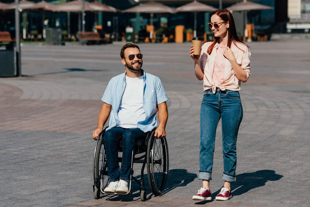 schöner Freund im Rollstuhl und Freundin mit Kaffee in Pappbecher beim Spazierengehen auf der Straße - Foto, Bild