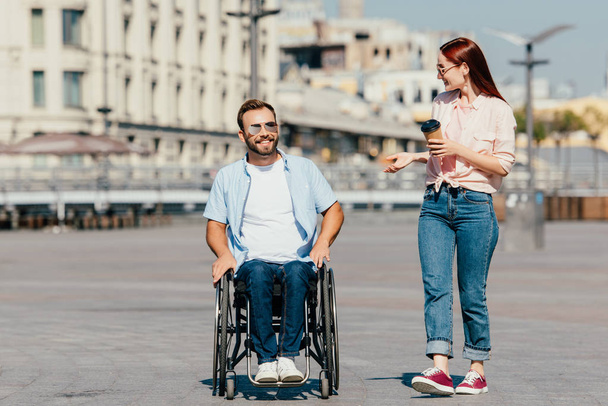 smiling handsome boyfriend in wheelchair and girlfriend with coffee to go having walk on street - Foto, immagini