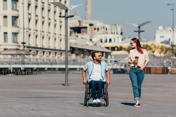 handsome boyfriend in wheelchair and girlfriend with having walk in city and looking at each other - Photo, Image
