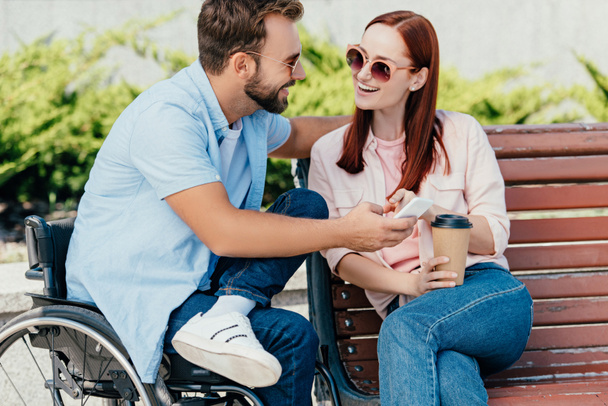 smiling handsome boyfriend in wheelchair and girlfriend holding smartphone and looking at each other on street - Foto, Imagen