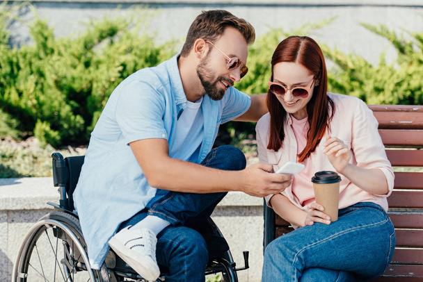 happy handsome boyfriend in wheelchair and girlfriend looking at smartphone on street - Photo, Image