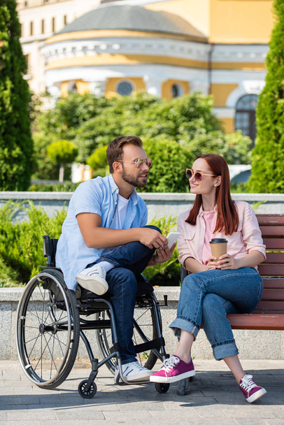 handsome boyfriend in wheelchair with smartphone and girlfriend with coffee to go on street - Фото, изображение
