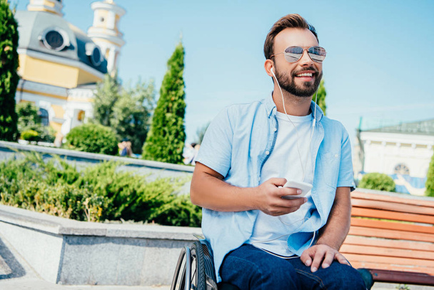 hombre guapo sonriente en silla de ruedas sosteniendo el teléfono inteligente y escuchando música en la calle, mirando hacia otro lado
 - Foto, Imagen