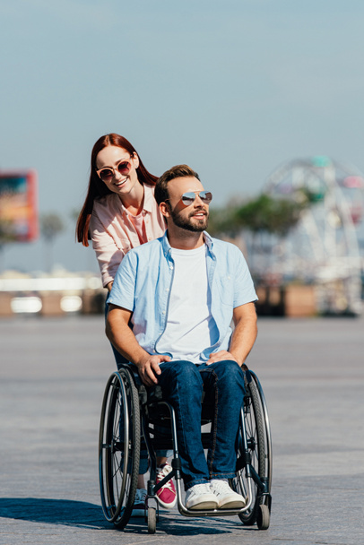 aantrekkelijke lachende vriendin knappe vriendje in rolstoel te duwen op straat - Foto, afbeelding