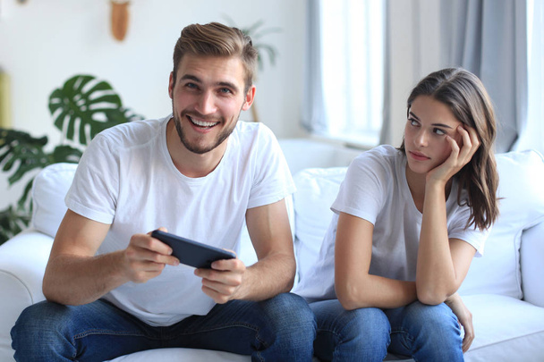 Man playing video games while his girlfriend is getting mad at him in their living room. - Zdjęcie, obraz