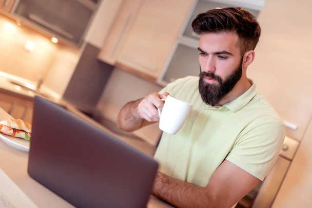 Счастливый человек работает на ноутбуке, пока пьет кофе на кухне
. - Фото, изображение