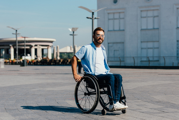 sonriente discapacitado guapo hombre en gafas de sol usando silla de ruedas en la calle
 - Foto, Imagen