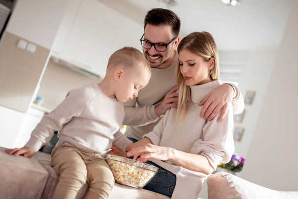 Onnellinen perhe äiti, isä ja poika istuu sohvalla kotona ja syö popcornia
. - Valokuva, kuva