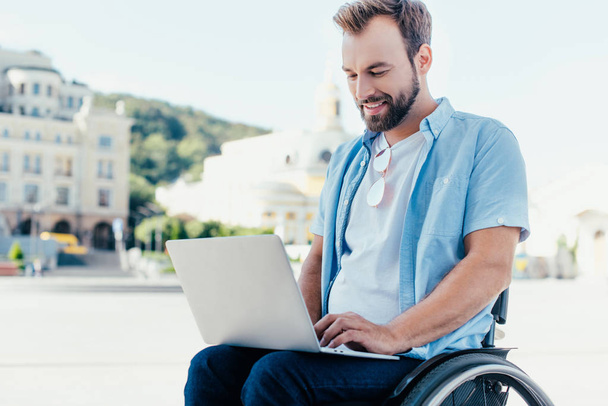 hombre guapo alegre en silla de ruedas usando el ordenador portátil en la calle
 - Foto, Imagen