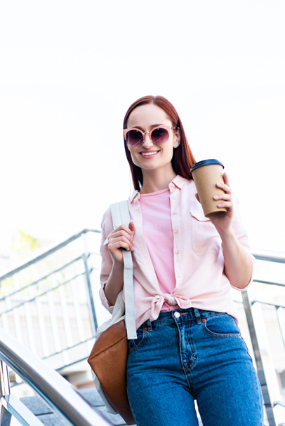 attractive redhair woman in pink shirt looking at camera on stairs with disposable coffee cup - Foto, imagen