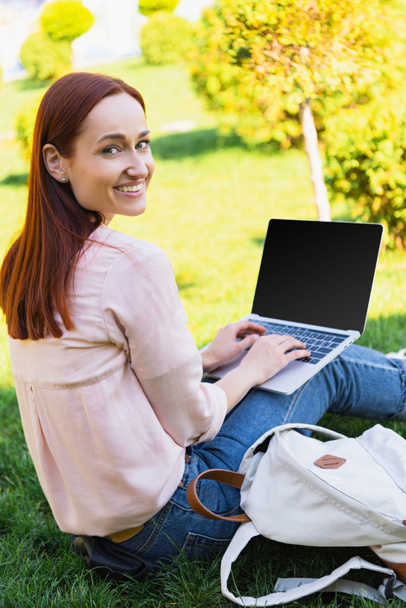 smiling attractive freelancer using laptop with blank screen in park and looking at camera - 写真・画像