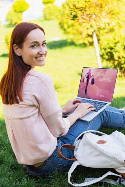 uśmiechający się atrakcyjną kobietę za pomocą laptopa z online zakupy urządzenia w parku i patrząc na kamery - Zdjęcie, obraz